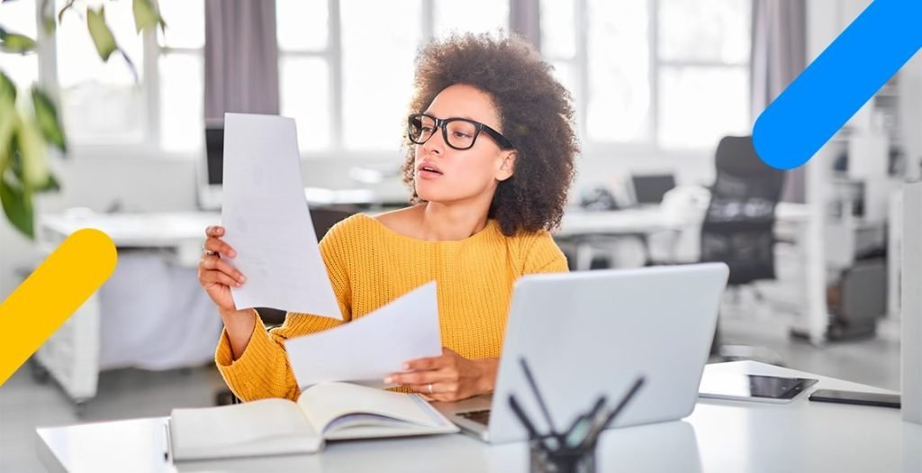 Mulher em frente a computador segurando e olhando para papéis nas mãos